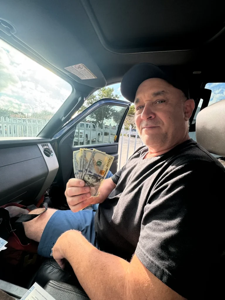 A man sitting in a vehicle holding a fan of $100 bills after selling his vehicle to a 