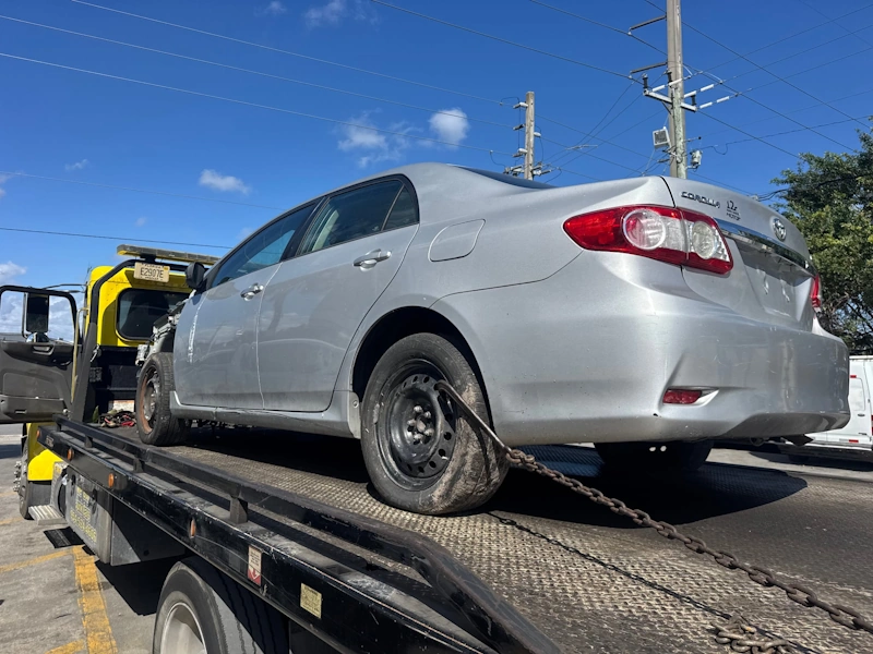 A silver Toyota Corolla being loaded onto a flatbed tow truck by a junk car buyer in Coconut Creek.