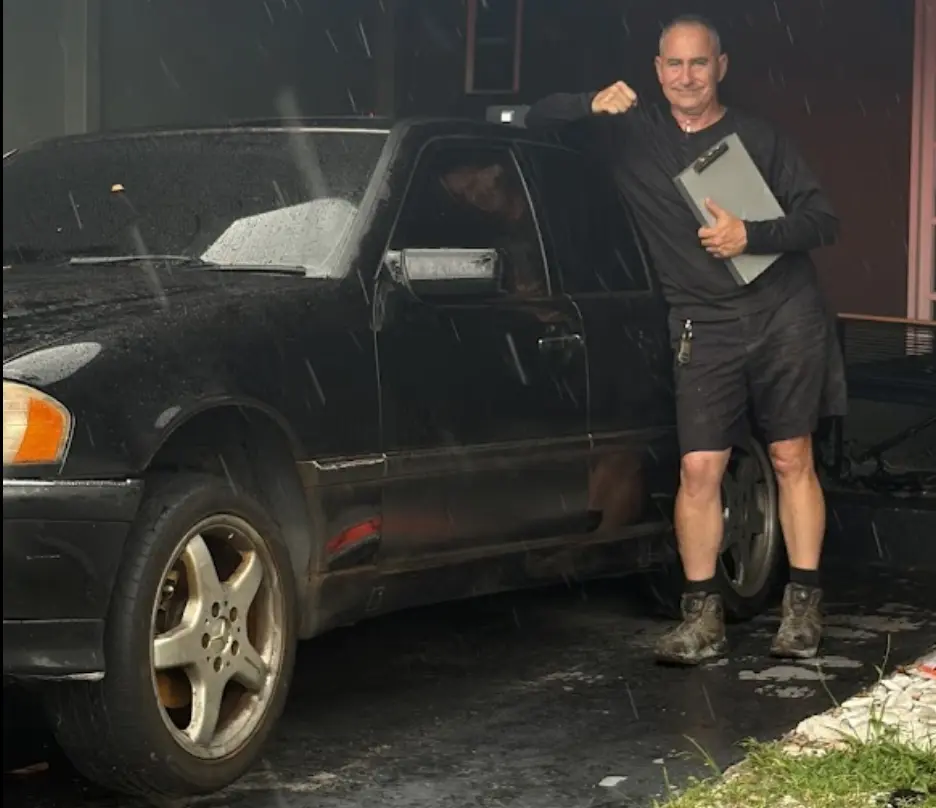 A professional buyer holding a clipboard stands next to a black sedan in the rain, demonstrating the reliable service offered by repeat junk car buyers in Tamarac.