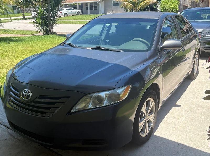 A worn, dark gray sedan parked on a residential concrete driveway, highlighting the hidden mechanical risks and dangers of driving a Junk Car.