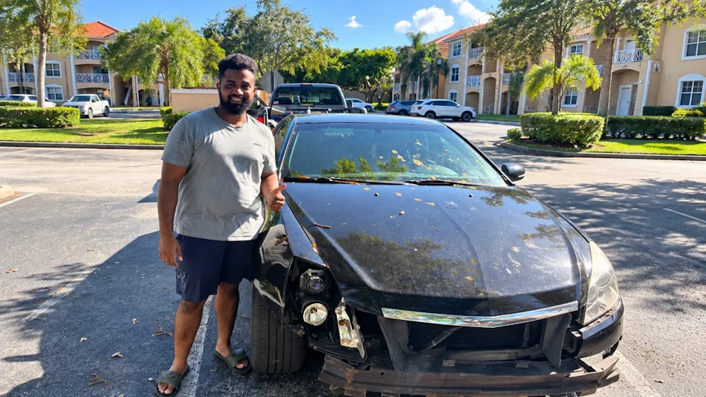 Smiling customer giving a thumbs up next to his wrecked Cadillac with a damaged front bumper and exposed headlight in Coconut Creek.